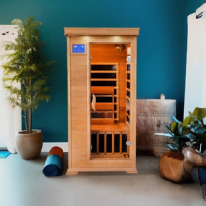 An indoor light-brown cedar infrared sauna stands against a bold teal-blue wall on a smooth gray floor. The sauna features a central glass door with a digital control panel on the upper left, a wooden bench seat inside, and dark heating panels. To the left is a tall potted plant and a rolled mat on the floor, and to the right sits a rustic wooden stump table and a leafy green plant.