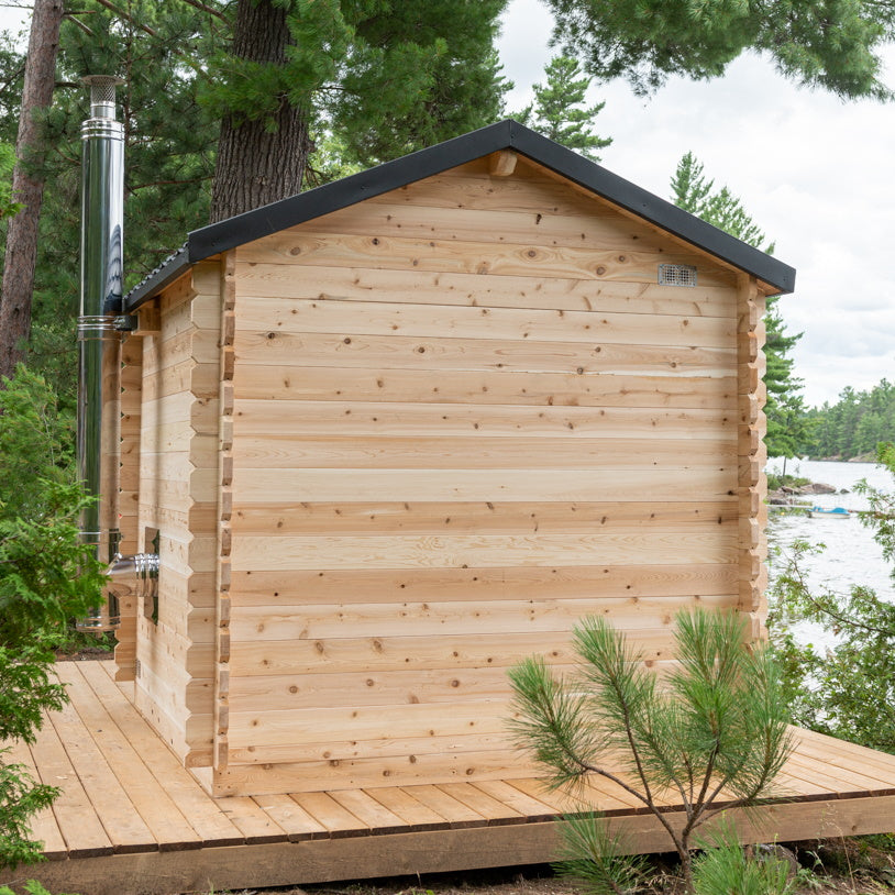 A small, freestanding log cabin sauna built from light, natural wood logs with interlocking corners. It sits on a wide wooden deck platform overlooking a large lake. The cabin features a peaked roof covered in dark grey, scalloped metal tiles, and a tall, vertical chrome chimney pipe extends up the side. The front has a central glass-paned door flanked by two narrow vertical windows. Tall pine trees grow alongside the deck on the right, under a cloudy sky.